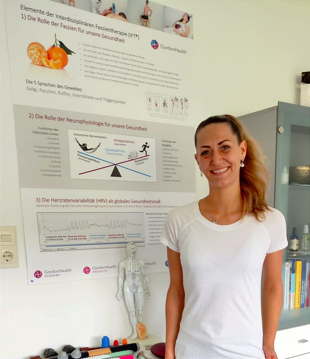 Maya in ihrer Praxis vor dem GordonHealth-Lehrposter zur Interdisziplinären Faszientherapie